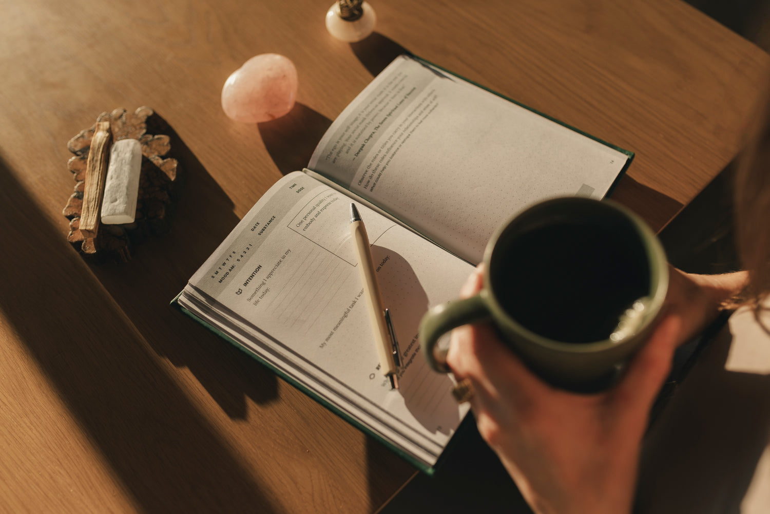 Open journal on a wood table beside a coffee mug, pen, and small crystals.