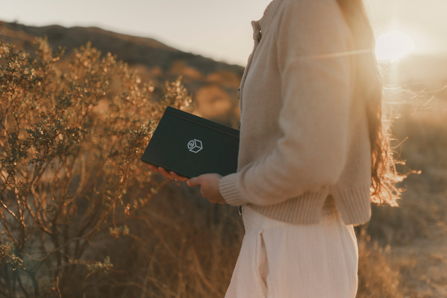 A woman walking through golden-hour brush, the closed journal at her hip.