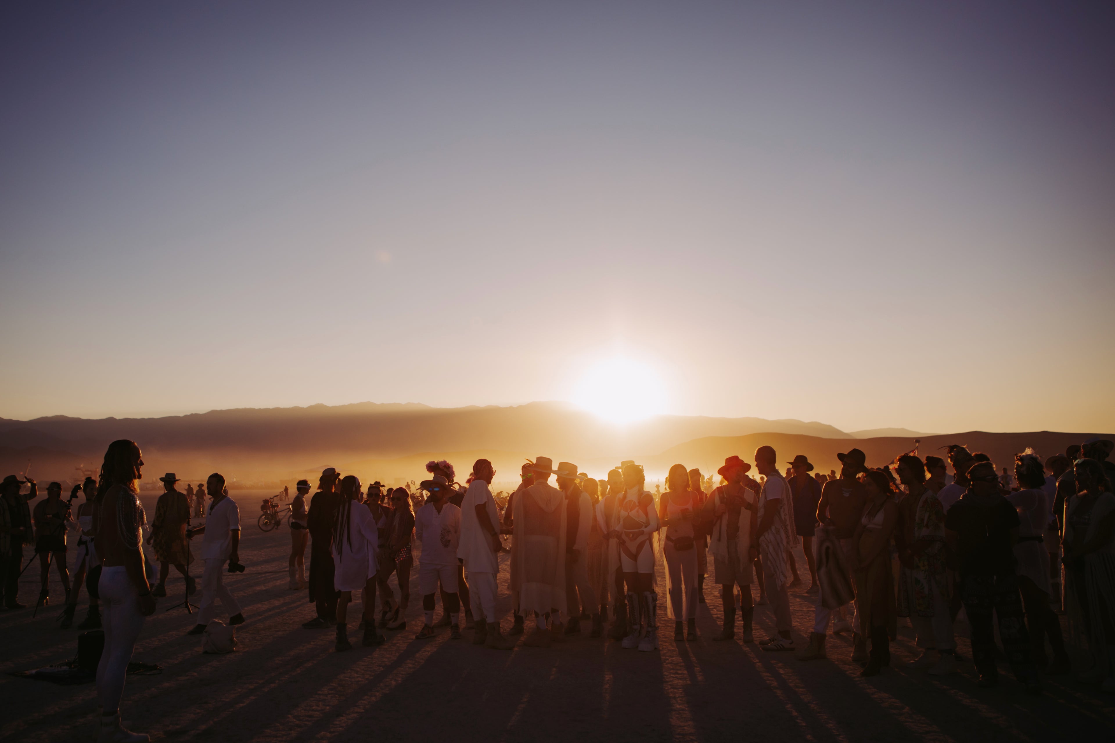 Silhouettes of a group of people gathered at sunset on the playa at Burning Man, with the mountains and golden light in the background, symbolizing community and connection.