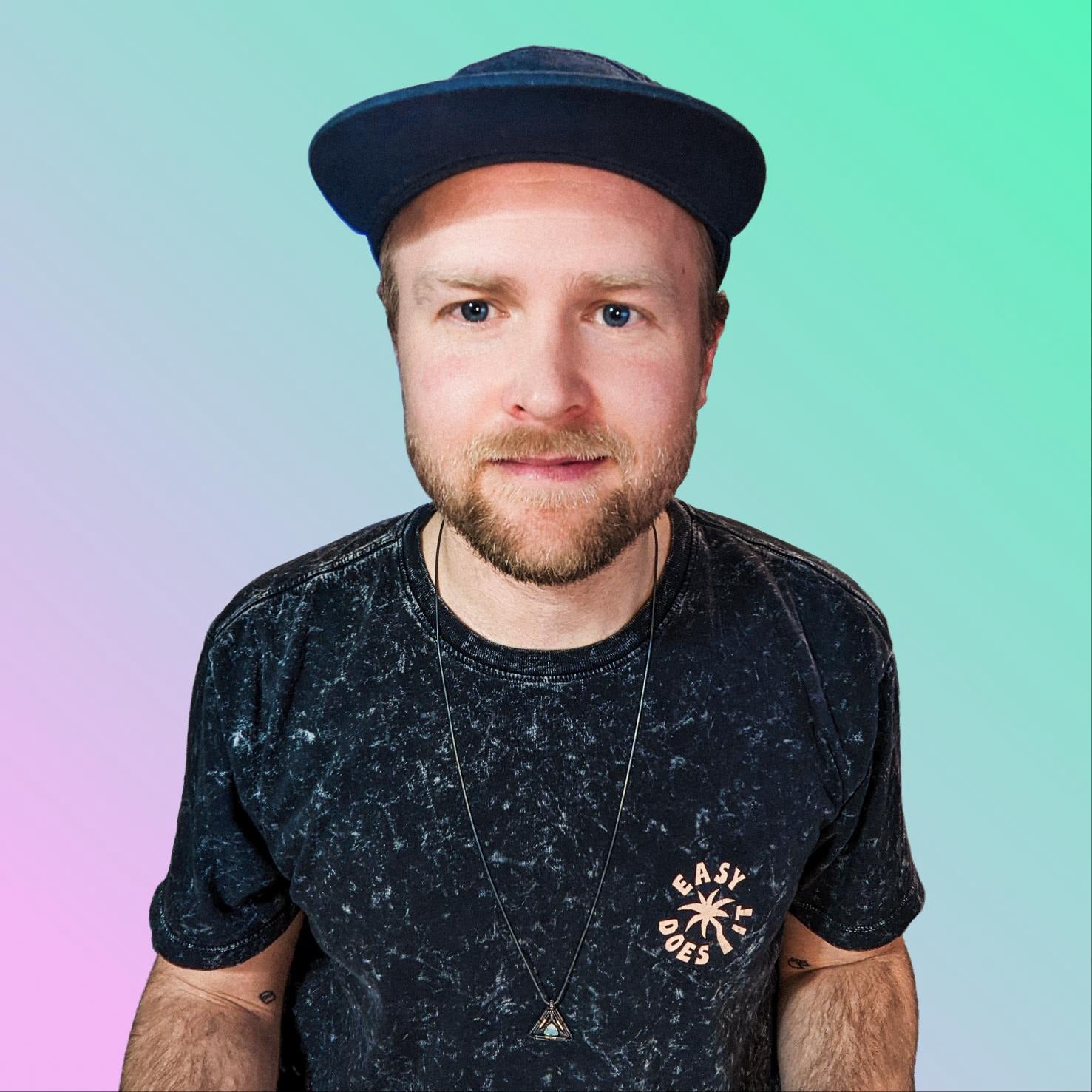 Cody Porter, Lead Designer of The Microdose Journal, wearing a dark patterned shirt and a triangular necklace, smiling against a pastel gradient background.